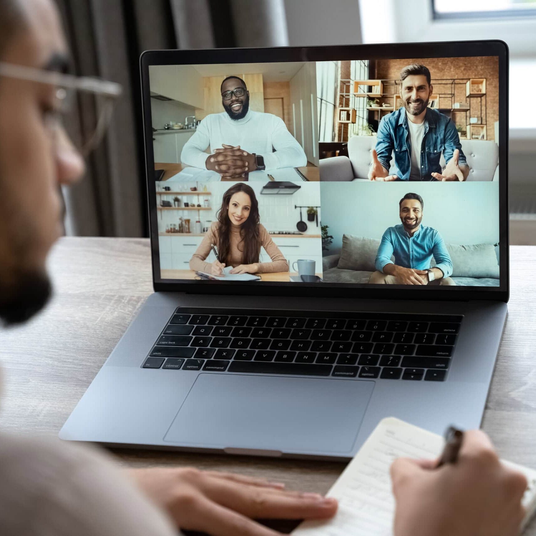 A person takes notes while participating in a video conference with four people displayed on a laptop screen.